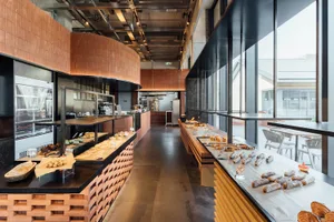 Modern bakery interior featuring a long curved counter with terracotta brick base, wooden display surfaces, various pastries and breads, large industrial windows, and concrete flooring.