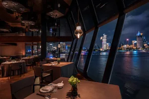 Upscale restaurant interior at night featuring dark wood beams, warm pendant lighting, linen-covered tables, and a panoramic view of the illuminated Shanghai skyline across the river.