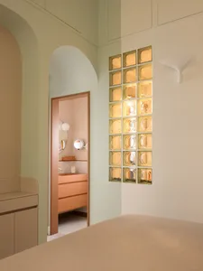Modern minimalist bedroom corner featuring sage green walls, a peach vanity visible through an arched doorway, illuminated yellow glass block wall, and a built-in beige storage bench.