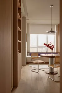 Modern minimalist dining nook with two cane and chrome chairs, round walnut table, built-in window seat with purple cushion, and large window overlooking trees.