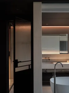 Modern minimalist bathroom featuring raw concrete walls, a white freestanding bathtub with a curved floor-mounted faucet, a stone floating vanity, and a black-framed ribbed glass door with warm backlighting.