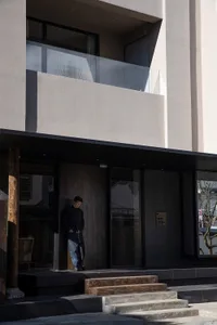 Modern minimalist house entrance featuring concrete walls, dark wood door and panels, wooden steps, and a man in black standing at the doorway.