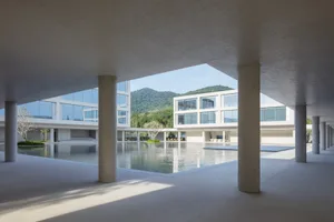 View from a shaded concrete colonnade overlooking a large rectangular reflecting pool, modern white buildings, and forested mountains in the background.