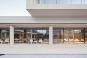 Modern minimalist concrete patio with cylindrical columns, white outdoor furniture, and large glass windows reflecting mountains at dusk.