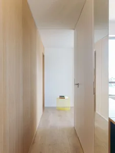 Narrow minimalist hallway lined with light oak wood paneling and cabinetry, featuring a bright yellow wireframe stool holding books at the end of the corridor.
