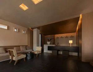 Modern minimalist lounge and reception area featuring exposed concrete walls, dark tiled floor, built-in beige sofa, woven cane armchairs, and a black reception counter with warm recessed lighting.