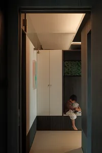 Modern minimalist entryway featuring dark wood paneling, white storage cabinets, a built-in bench, and a young boy putting on shoes under soft overhead lighting.
