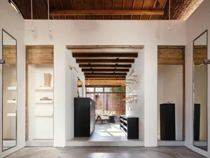 Modern minimalist boutique interior featuring white walls, black display fixtures, and prominent reclaimed wooden ceiling beams with natural light from rear courtyard.
