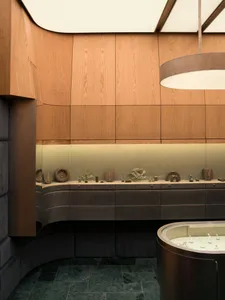 Interior view of a luxury jewelry store featuring curved oak wood paneling, dark green marble floor tiles, integrated backlit display cases, and a large circular pendant light hanging above a central curved glass display counter.