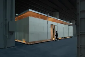 Modern concrete modular pavilion with warm wooden ceiling, integrated LED lighting at the base, and a person walking past to show scale, inside a large industrial hall.