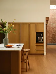 Modern minimalist kitchen featuring a curved oak island with white marble countertop, wooden stool, built-in oak cabinetry, and a large grey vase with yellow forsythia branches.