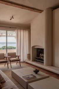 Modern Mediterranean living room featuring a curved plaster fireplace, terracotta floor tiles, wooden beam ceiling, two wooden armchairs, and large glass doors opening to a sea view.