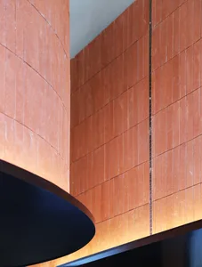 Close-up view of a curved terracotta tile wall meeting a flat terracotta tile wall, with warm hidden lighting glowing from below a dark circular architectural element.