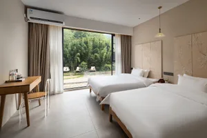 Modern twin bed hotel room with white bedding, floral pattern headboards, wooden desk, and large glass doors opening to a green bamboo forest view.