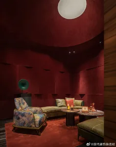 Modern lounge with deep red curved walls, patterned armchair, green velvet sofa, round marble coffee table, and large spherical pendant light.