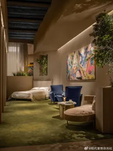Modern bedroom interior with olive-green carpet, two sapphire blue velvet armchairs, a round woven ottoman, large colorful abstract painting, and a low platform bed with white linens.