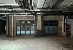 Empty raw industrial loft interior with exposed concrete pillars, brick walls, overhead black piping and ductwork, and large glass doors.