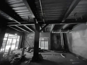 Black and white photograph of an unfinished industrial loft interior showing exposed steel beams, concrete ceiling, structural columns, and large windows with construction debris on the floor.