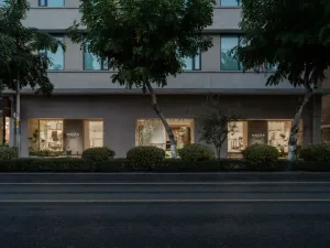 Evening view of a minimalist concrete retail building with warm interior lighting, large glass openings, and trees lining the street.