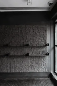 Monochrome interior featuring a textured stacked stone wall with three black floating shelves, dark tiled floor, and industrial-style lighting fixtures.