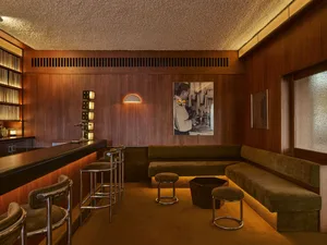 Mid-century modern home bar interior with walnut wood walls, olive green velvet L-shaped sofa and bar stools, chrome accents, and warm ambient lighting.