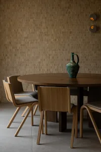 Modern minimalist dining area with a round oak table, four matching oak dining chairs with grey upholstery, a green ceramic pitcher, and a warm brown textured tile wall with two circular wall sconces.