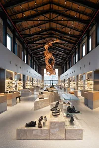 Long view down a modern shoe store with travertine display blocks, wooden floating sculpture, industrial ceiling, and extensive shelving.