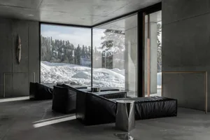 現代極簡客廳空間，原始混凝土牆面、黑色皮革模組沙發、金屬邊桌，以及落地大窗俯瞰覆雪山林。