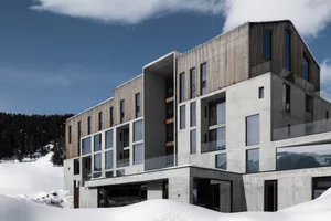 Modern multi-story alpine resort building featuring exposed concrete, vertical timber slats, large windows, and glass balconies, set in a snowy mountain environment under a clear blue sky.