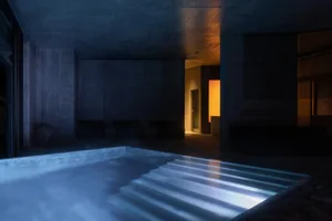 Night view of an indoor swimming pool in a brutalist concrete structure with dramatic blue underwater lighting and warm orange accent lighting in the background corridor.