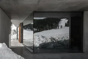 Interior view of a modern concrete mountain house looking out onto a snow-covered alpine forest and ski slope.