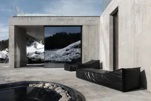 Modern concrete villa exterior featuring black leather outdoor lounge beds, a circular hot tub with river stones, and a large glass window reflecting a snowy forest and mountain.