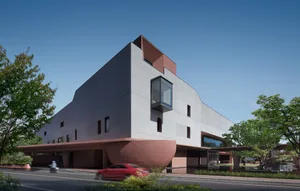 Exterior view of a modern Brutalist-style cultural center building with grey concrete upper volumes and a curved terracotta-colored base, surrounded by green trees under a clear blue sky.