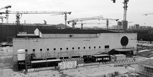 Black and white photograph of a large concrete industrial building with a circular window, surrounded by multiple construction cranes at a demolition and redevelopment site.