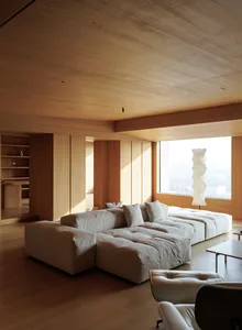 Modern minimalist living room featuring a large low-profile modular sofa in off-white fabric, extensive light oak wood paneling on walls and ceiling, a sculptural paper floor lamp, and leather lounge chairs in the foreground.