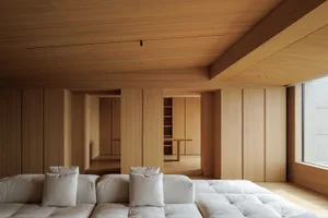 Modern minimalist living room featuring a large modular white sofa, extensive oak built-in cabinetry, wooden ceiling, and a view into a dining area.