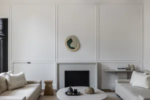 Minimalist living room with two cream sofas facing a round concrete coffee table, a black fireplace with limestone surround, and an irregular organic mirror on a white paneled wall.