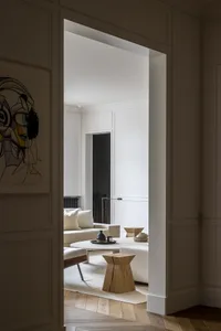 View through a doorway into a minimalist living room featuring a cream linen sofa, sculptural oak side tables, a round travertine coffee table, and a large abstract portrait painting.