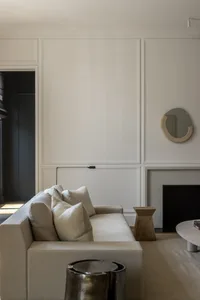 Modern neutral living room featuring a beige linen sofa with plush pillows, a sculptural oak side table, a metallic coffee table, and an irregular organic mirror against white panelled walls.