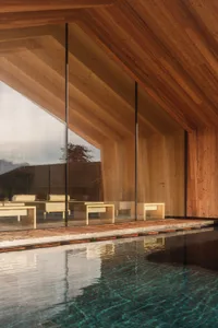 Modern indoor swimming pool with clear turquoise water, surrounded by warm teak wood walls and ceiling, large glass windows overlooking mountains, and minimalist wooden lounge furniture visible through the glass.