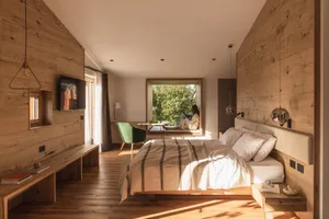 Modern minimalist bedroom with extensive light wood paneling, a large bed with striped linen, built-in window seat with a woman sitting, green velvet chair, and pendant lights.