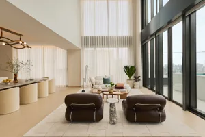 Modern minimalist living room featuring two brown leather armchairs, a marble side table, a light wood coffee table, and a white sofa in the background, with floor-to-ceiling windows overlooking a city skyline.