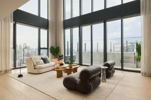 Modern luxury living room featuring a brown leather chaise lounge, cream fabric sofa, wooden coffee tables, and expansive floor-to-ceiling windows overlooking a dense city skyline.