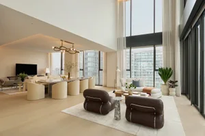 Modern luxury living room featuring two brown leather lounge chairs in the foreground, a large marble dining table with cream boucle chairs, high ceilings, and expansive city views through full-height windows.
