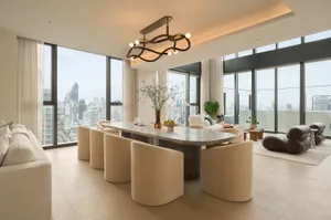 Modern open-plan dining area featuring a large curved marble table surrounded by cream boucle chairs, with floor-to-ceiling windows showcasing a dense city skyline.