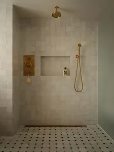 Modern luxury shower corner with light marble tiled walls, recessed niche, unlacquered brass shower head and valve, and geometric black and white marble floor tiles.