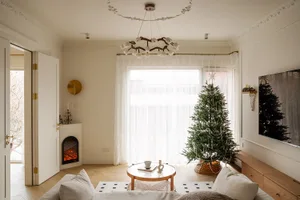 Bright living room with white sofa, round glass coffee table, decorated Christmas tree by the window, electric fireplace, and oak TV console.