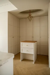 Modern classic dressing room with cream built-in wardrobes, oak island cabinet, herringbone wood floor and ornate crystal chandelier hanging from a circular ceiling detail.