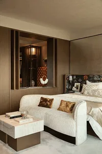 Modern luxury bedroom interior with a curved white bouclé sofa, two gold velvet pillows, marble coffee table with books, walnut wood paneling, and a floral patterned headboard on the bed.