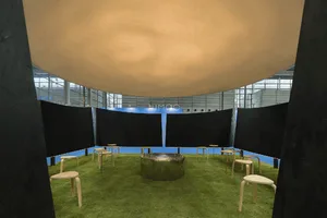 Modern art installation showing a large beige fabric dome suspended above a circular green artificial grass floor with wooden stools and a central metallic fire pit.